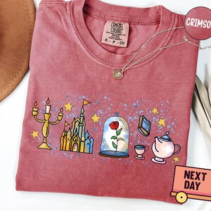 May include: A crimson red t-shirt with a graphic print of a castle, a rose in a glass dome, a candlestick, a book, and a teapot. The text "COMFORT COLORS" is printed on the shirt.