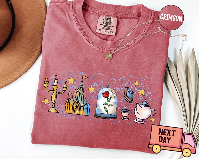 May include: A crimson red t-shirt with a graphic print of a castle, a rose in a glass dome, a candlestick, a book, and a teapot. The text "COMFORT COLORS" is printed on the shirt.