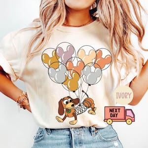 May include: A white t-shirt with a cartoon image of a dog holding a bunch of balloons shaped like Mickey Mouse ears. The balloons are in shades of pink, orange, yellow, and gray. The text "IVORY" and "NEXT DAY" are visible on the image.