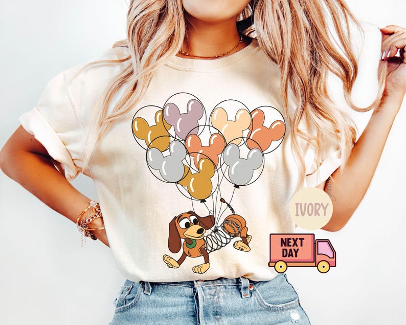 May include: A white t-shirt with a cartoon image of a dog holding a bunch of balloons shaped like Mickey Mouse ears. The balloons are in shades of pink, orange, yellow, and gray. The text "IVORY" and "NEXT DAY" are visible on the image.