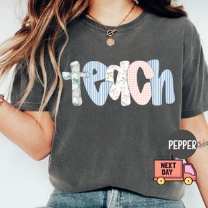 May include: Dark gray t-shirt with the word "teach" in a patchwork design. The letters are in floral, gingham, and striped patterns. The shirt has a crew neck and short sleeves.