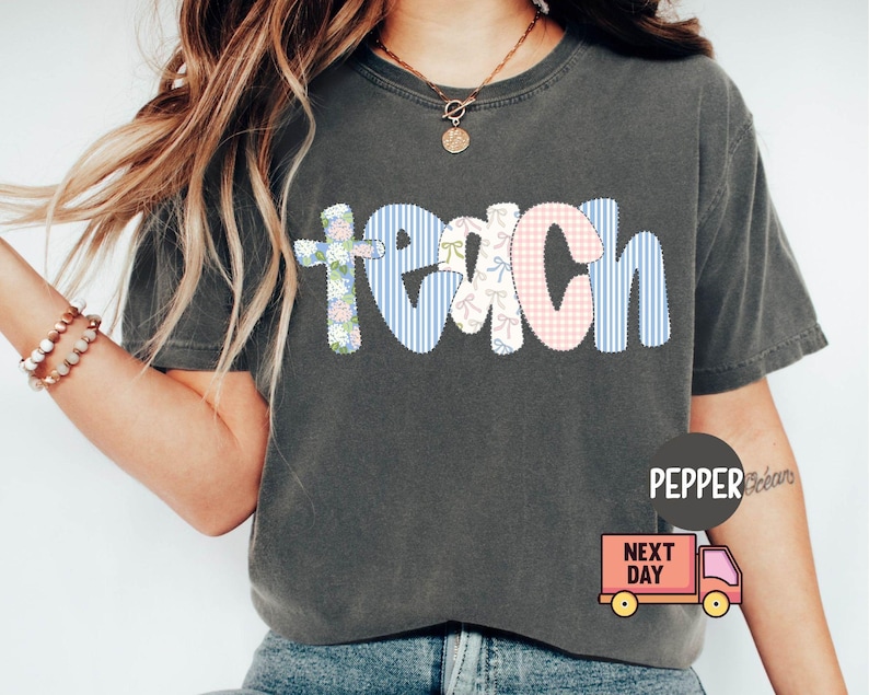May include: Dark gray t-shirt with the word "teach" in a patchwork design. The letters are in floral, gingham, and striped patterns. The shirt has a crew neck and short sleeves.