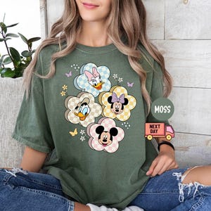 May include: Green t-shirt featuring a floral design with cartoon characters. The flowers have a checkered pattern and feature Mickey Mouse, Minnie Mouse, Donald Duck, and Daisy Duck. The shirt has short sleeves and a relaxed fit.