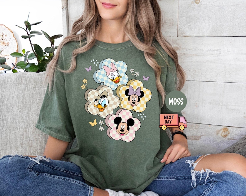 May include: Green t-shirt featuring a floral design with cartoon characters. The flowers have a checkered pattern and feature Mickey Mouse, Minnie Mouse, Donald Duck, and Daisy Duck. The shirt has short sleeves and a relaxed fit.