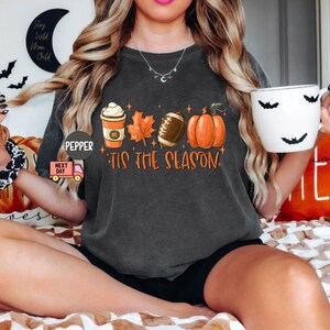 May include: A dark gray t-shirt with a graphic of a coffee cup, a football, a pumpkin, and leaves. The text "Tis the season" is printed on the shirt.