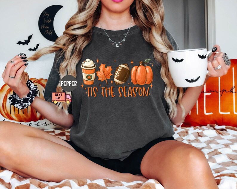 May include: A dark gray t-shirt with a graphic of a coffee cup, a football, a pumpkin, and leaves. The text "Tis the season" is printed on the shirt.