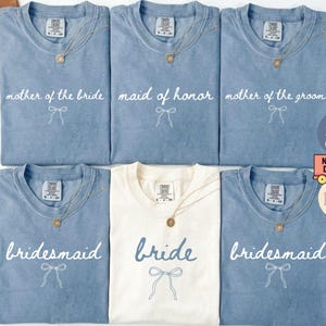 May include: A selection of blue jean and ivory t-shirts with wedding-related text. The shirts feature phrases such as "mother of the bride," "maid of honour," and "bride." Each shirt has a bow graphic and a gold necklace.