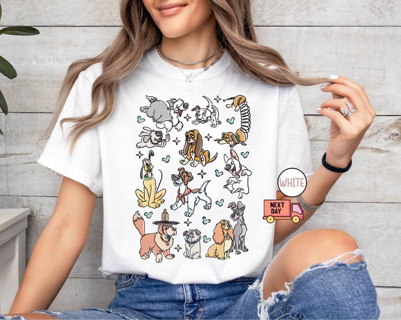 May include: White t-shirt featuring a colorful cartoon illustration of various dog breeds. The design includes dogs of different sizes and colors, with a playful and whimsical style. The shirt has a round neckline and short sleeves.