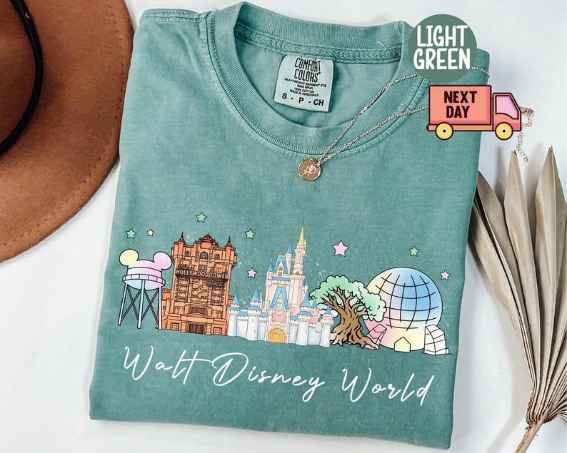 May include: Light green t-shirt with a Walt Disney World graphic. The design includes a water tower, Hollywood Tower, Cinderella Castle, and a Spaceship Earth illustration. The words "Walt Disney World" are written in cursive.