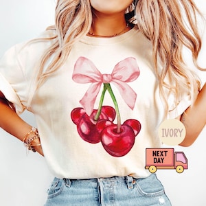 Puede incluir: Camiseta color marfil con un dise&ntilde;o de acuarela de tres cerezas rojas y un lazo rosa. La camiseta tiene mangas cortas y un corte relajado. El texto "IVORY" est&aacute; impreso en la camiseta.