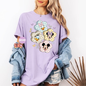 May include: A light purple t-shirt featuring a floral design with cartoon characters Daisy Duck, Donald Duck, Minnie Mouse, and Mickey Mouse. The flowers have a checkered pattern in pastel colors. The shirt has short sleeves and a relaxed fit.