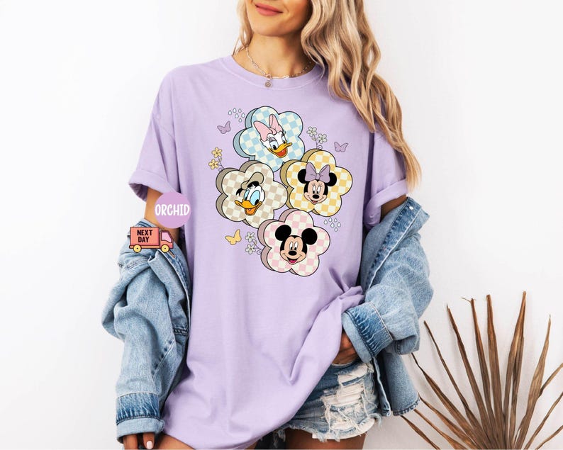 May include: A light purple t-shirt featuring a floral design with cartoon characters Daisy Duck, Donald Duck, Minnie Mouse, and Mickey Mouse. The flowers have a checkered pattern in pastel colors. The shirt has short sleeves and a relaxed fit.