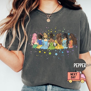 Puede incluir: Una camiseta gris oscuro con un gr&aacute;fico de siete mujeres con cabello largo, vestidas con vestidos coloridos y sentadas de espaldas. El gr&aacute;fico est&aacute; rodeado de estrellas.