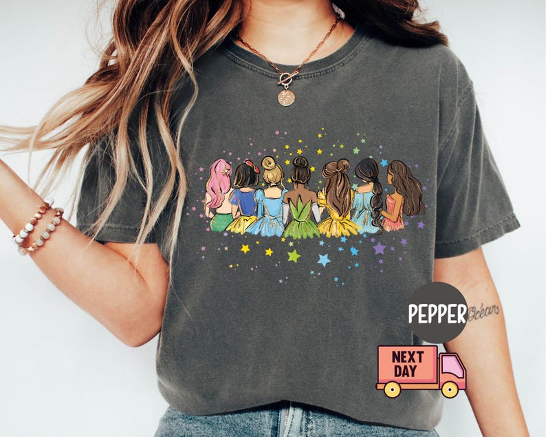 Puede incluir: Una camiseta gris oscuro con un gr&aacute;fico de siete mujeres con cabello largo, vestidas con vestidos coloridos y sentadas de espaldas. El gr&aacute;fico est&aacute; rodeado de estrellas.