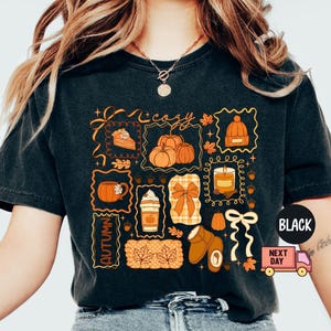 May include: Black t-shirt with a colorful autumn-themed graphic. The design includes pumpkins, a slice of pie, a mug of coffee, a candle, and the word "cozy" and "autumn". The shirt has a vintage, washed look.