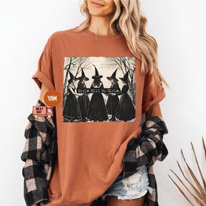 May include: A rust-colored t-shirt with a graphic of three witches wearing black pointed hats and robes. The text "Girls Will Be Girls" is printed on the shirt.