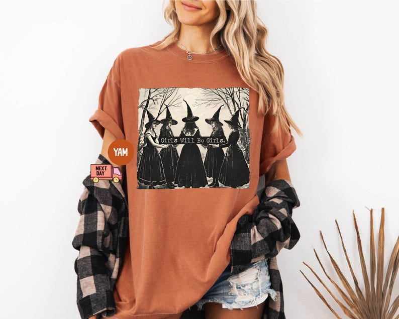 May include: A rust-colored t-shirt with a graphic of three witches wearing black pointed hats and robes. The text "Girls Will Be Girls" is printed on the shirt.