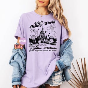 May include: A light purple t-shirt featuring a black graphic of the Walt Disney World castle, balloons, and fireworks. The text "Walt Disney World" and "The happiest place on earth" are printed on the shirt.