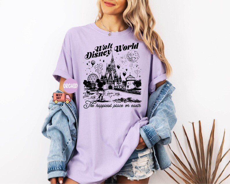 May include: A light purple t-shirt featuring a black graphic of the Walt Disney World castle, balloons, and fireworks. The text "Walt Disney World" and "The happiest place on earth" are printed on the shirt.