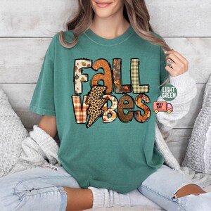 May include: A teal blue t-shirt featuring the text "Fall Vibes" in a patchwork style with autumnal colours and patterns. The text is in a bold, playful font.