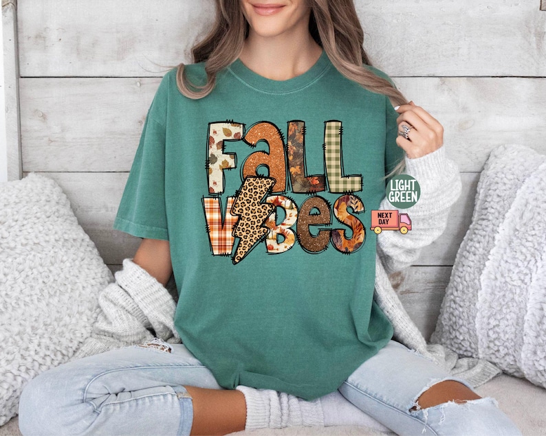 May include: A teal blue t-shirt featuring the text "Fall Vibes" in a patchwork style with autumnal colours and patterns. The text is in a bold, playful font.