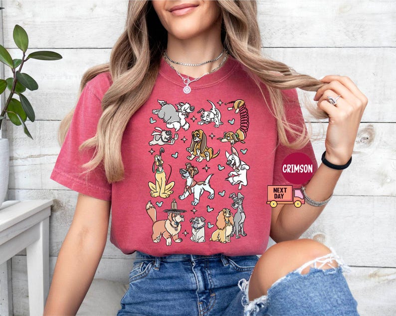 May include: A red t-shirt featuring a collection of cartoon dogs in various poses. The dogs are in different colors and breeds, with hearts and stars scattered around them. The shirt has the text "CRIMSON" and "NEXT DAY" on it.