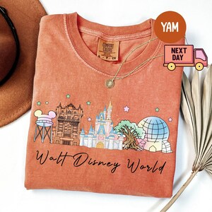 May include: Coral-colored t-shirt featuring a Walt Disney World graphic. The design includes iconic park landmarks like Cinderella Castle and Spaceship Earth, with the words "Walt Disney World" in script. A gold necklace and a brown hat are also visible.