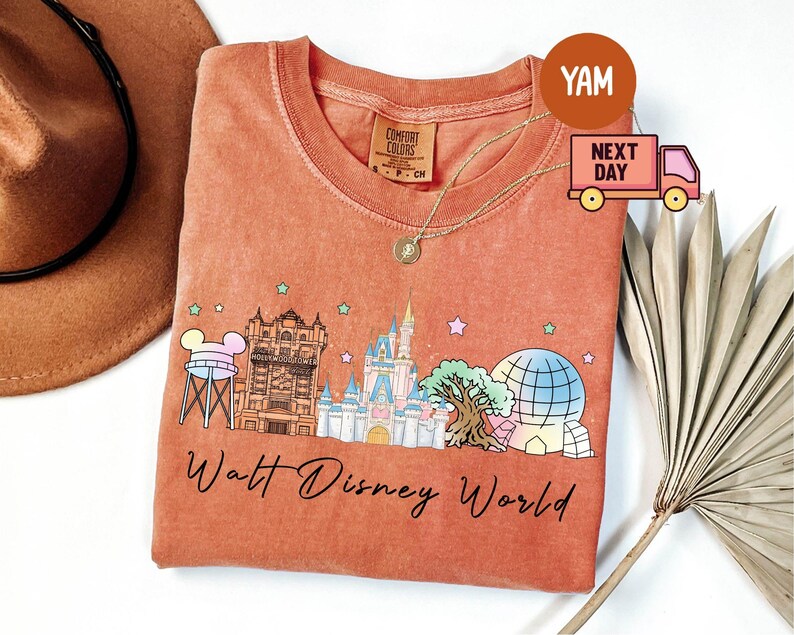 May include: Coral-colored t-shirt featuring a Walt Disney World graphic. The design includes iconic park landmarks like Cinderella Castle and Spaceship Earth, with the words "Walt Disney World" in script. A gold necklace and a brown hat are also visible.