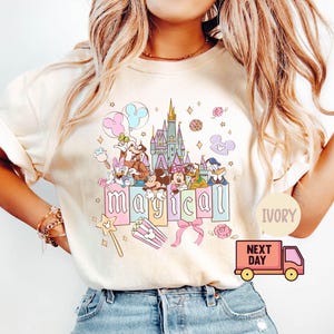 May include: Ivory-colored t-shirt featuring a colorful graphic of Disney characters, a castle, and the word "magical." The design includes Mickey and Minnie Mouse, Donald Duck, and other characters. Pink and blue balloons and a pink bow are also included.