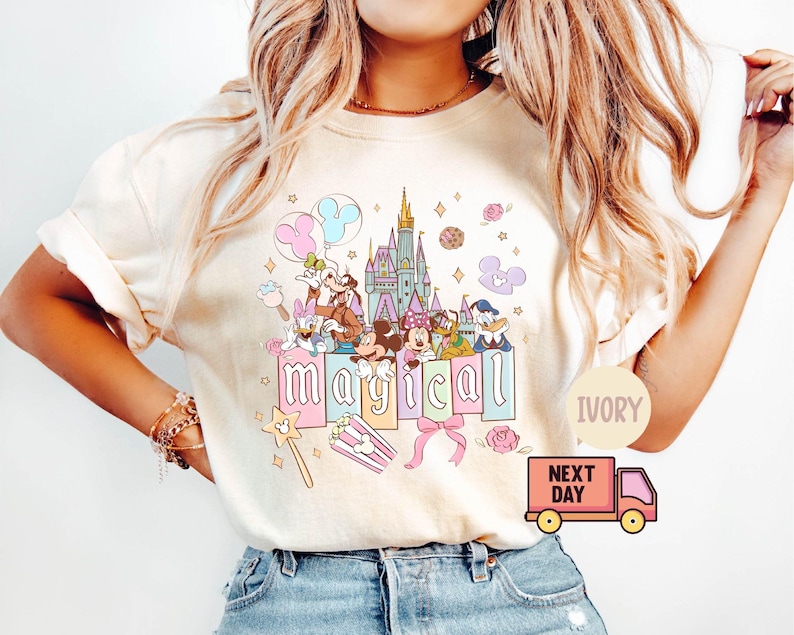May include: Ivory-colored t-shirt featuring a colorful graphic of Disney characters, a castle, and the word "magical." The design includes Mickey and Minnie Mouse, Donald Duck, and other characters. Pink and blue balloons and a pink bow are also included.