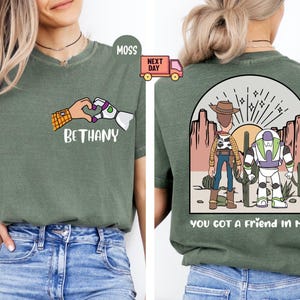 Pu&ograve; includere: Una t-shirt verde muschio con una grafica frontale che mostra un cuore formato dalle mani di un cowboy e di un ranger spaziale con il nome BETHANY. Il retro mostra i due personaggi in un paesaggio desertico con il testo YOU GOT A FRIEND IN ME.