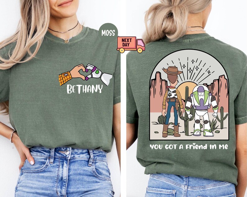 Pu&ograve; includere: Una t-shirt verde muschio con una grafica frontale che mostra un cuore formato dalle mani di un cowboy e di un ranger spaziale con il nome BETHANY. Il retro mostra i due personaggi in un paesaggio desertico con il testo YOU GOT A FRIEND IN ME.