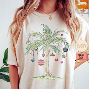 Pode incluir: Uma camiseta branca com um design de palmeira e ornamentos coloridos. O texto "IVORY" e "NEXT DAY" é visível na camiseta.