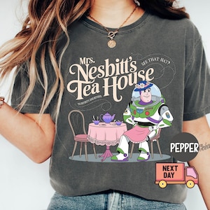 May include: Dark gray t-shirt featuring a cartoon illustration of Buzz Lightyear as Mrs. Nesbitt from Toy Story. The design includes a tea table, text that reads "Mrs. Nesbitt's Tea House", and the phrase "To Infinity and Beyond".