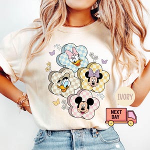May include: Ivory-colored t-shirt featuring a floral design with cartoon characters. The flowers have checkered patterns and feature Daisy Duck, Donald Duck, Minnie Mouse, and Mickey Mouse. The shirt has short sleeves and a round neckline.