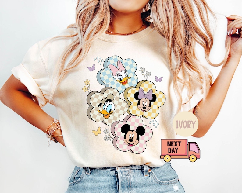 May include: Ivory-colored t-shirt featuring a floral design with cartoon characters. The flowers have checkered patterns and feature Daisy Duck, Donald Duck, Minnie Mouse, and Mickey Mouse. The shirt has short sleeves and a round neckline.