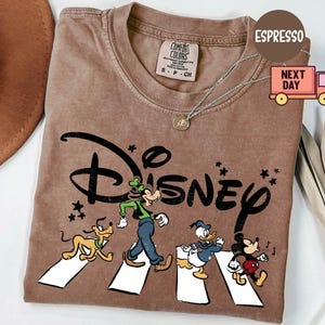 K&ouml;nnte beinhalten: Espressofarbenes T-Shirt mit dem Disney-Logo und Zeichentrickfiguren, die eine Stra&szlig;e &uuml;berqueren. Das Design umfasst Pluto, Goofy, Donald Duck und Mickey Mouse. Das Shirt hat einen Rundhalsausschnitt und kurze &Auml;rmel.