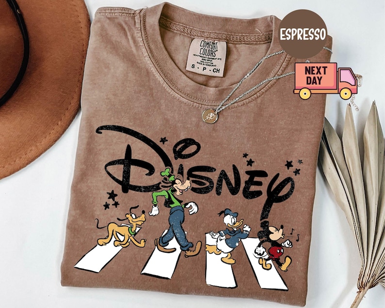 K&ouml;nnte beinhalten: Espressofarbenes T-Shirt mit dem Disney-Logo und Zeichentrickfiguren, die eine Stra&szlig;e &uuml;berqueren. Das Design umfasst Pluto, Goofy, Donald Duck und Mickey Mouse. Das Shirt hat einen Rundhalsausschnitt und kurze &Auml;rmel.