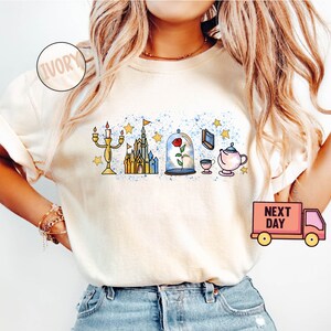 May include: A white t-shirt with a graphic design featuring characters from the Disney movie Beauty and the Beast. The design includes a castle, a rose in a glass dome, a candlestick, a teapot, and a teacup. The shirt is available in ivory.