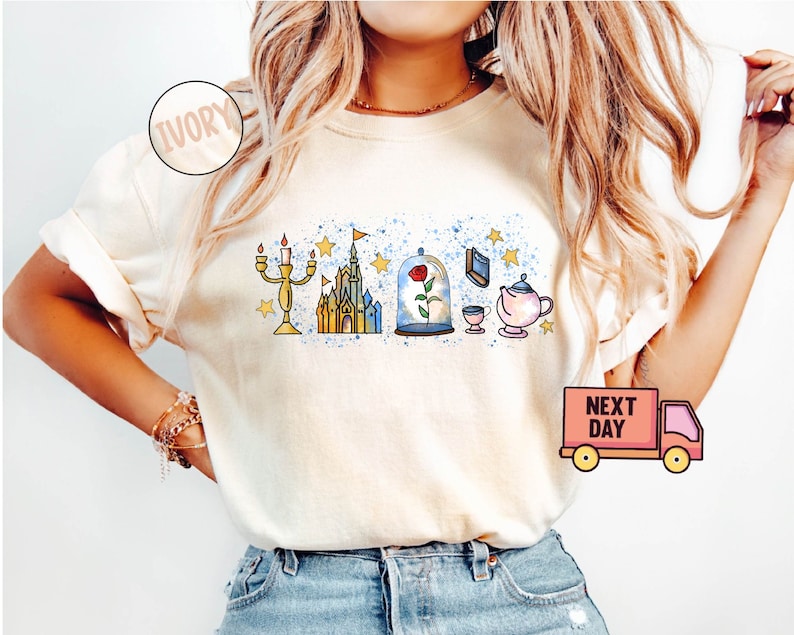 May include: A white t-shirt with a graphic design featuring characters from the Disney movie Beauty and the Beast. The design includes a castle, a rose in a glass dome, a candlestick, a teapot, and a teacup. The shirt is available in ivory.