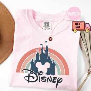 May include: Pink t-shirt with a rainbow and a silhouette of Cinderella's castle with Mickey Mouse ears and the word "DISNEY" in a stylized font.