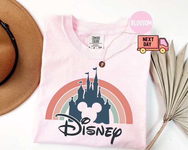May include: Pink t-shirt with a rainbow and a silhouette of Cinderella's castle with Mickey Mouse ears and the word "DISNEY" in a stylized font.