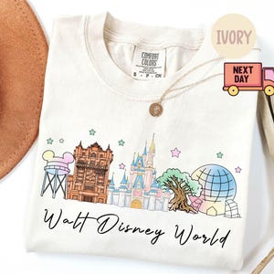 May include: Ivory-colored crew neck t-shirt featuring a colorful illustration of Walt Disney World landmarks. The design includes the Tower of Terror, Cinderella Castle, and a Spaceship Earth-like structure, with the words "Walt Disney World" in script.
