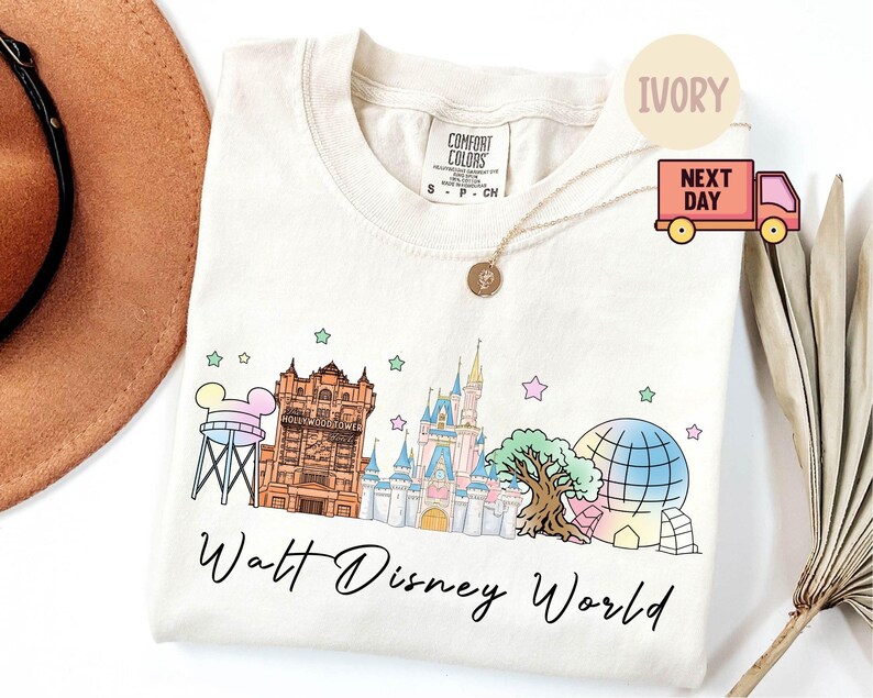 May include: Ivory-colored crew neck t-shirt featuring a colorful illustration of Walt Disney World landmarks. The design includes the Tower of Terror, Cinderella Castle, and a Spaceship Earth-like structure, with the words "Walt Disney World" in script.