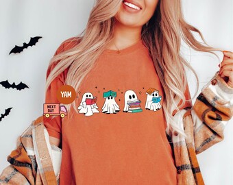 Op de afbeelding: Oranje T-shirt met een afbeelding van vier geesten die boeken lezen. De geesten zijn wit met zwarte ogen. De boeken zijn kleurrijk en op elkaar gestapeld. Het shirt heeft een ronde hals en korte mouwen.