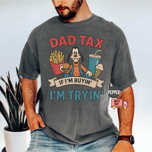 Peut inclure: T-shirt gris avec un personnage de dessin animé tenant un hamburger et des frites, avec le texte "DAD TAX" au-dessus et "IF I'M BUYIN' I'M TRYIN" en dessous. Le t-shirt a un aspect vintage.
