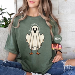 May include: Moss green t-shirt featuring a cartoon ghost with goose feet and the text "Just a Spooky Goose". The ghost is white with black eyes and a beak. The shirt is worn by a person.