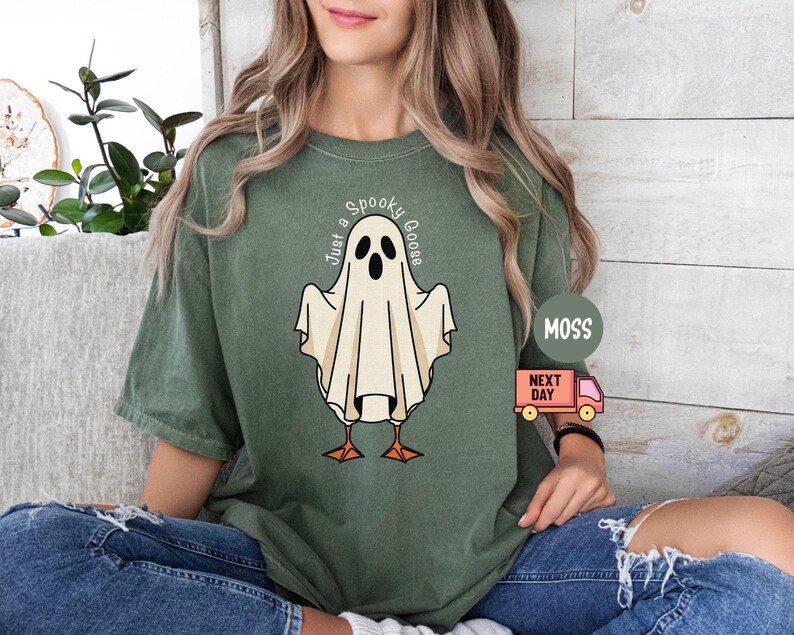 May include: Moss green t-shirt featuring a cartoon ghost with goose feet and the text "Just a Spooky Goose". The ghost is white with black eyes and a beak. The shirt is worn by a person.