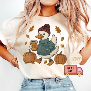 May include: Ivory t-shirt with an embroidered duck wearing a hat, scarf, and boots, holding a coffee cup. Two pumpkins and autumn leaves surround the duck. The shirt has the word "IVORY" printed on it.