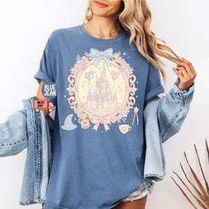 K&ouml;nnte beinhalten: Ein hellblaues T-Shirt mit einer pastellrosa und blauen Illustration von Schloss Aschenputtel, Mickey-Maus-Ohren und einer Teetasse. Die Illustration ist von einem Blumengirlanden und einer Schleife umgeben.